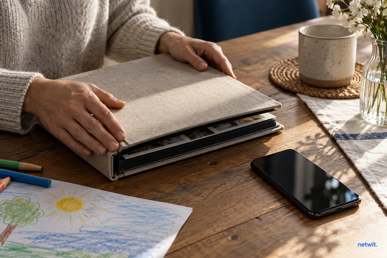 An adult's hands closing a family photo album beside a face-down smartphone, a child's small drawing partially visible, no child faces.